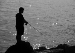 きらめく海を前に岩場で竿を構える人のシルエット